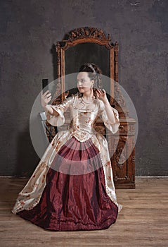 Beautiful woman in rococo style medieval dress sitting near console mirror table with mobile phone making selfie photo eclecticism