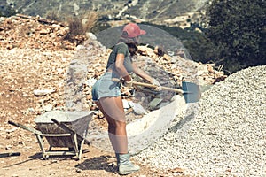 Beautiful woman in the shorts and in the red peaked cap with a wheelbarrow for cement and the shovel digging on stone. Concept