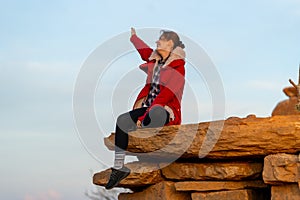 Beautiful woman sit on the rock near cliff on the mountain and raise her arm up with happiness and look forward with warm light in