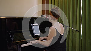 Beautiful woman sits down and starts to play the piano while looking at the tablet