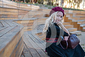 A beautiful woman sits on a wooden summer theater bench in a burgundy coat and biret, an adult smiles at the camera, in