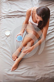 Beautiful woman sitting on bed and applying cream on legs