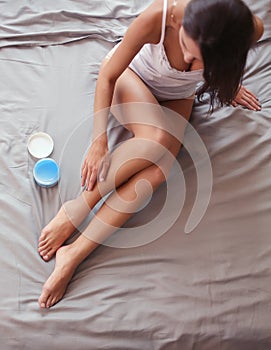 Beautiful woman sitting on bed and applying cream on legs