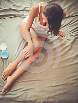 Beautiful woman sitting on bed and applying cream on legs