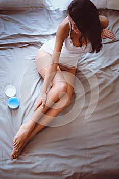 Beautiful woman sitting on bed and applying cream on legs