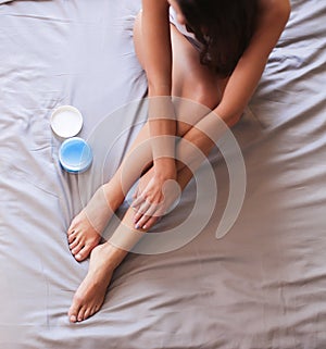 Beautiful woman sitting on bed and applying cream on legs