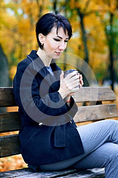 Beautiful woman sitting with coffee and dreaming in autumn city park, fall season, yellow leaves