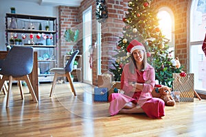 Beautiful woman sitting on the floor around christmas tree and presents wearing santa claus hat and smiling happy