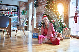 Beautiful woman sitting on the floor around christmas tree and presents wearing santa claus hat and smiling happy