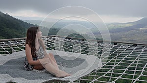 Beautiful woman sitting on a hammock net couch outdoors above green misty abyss. Korean female tourist chilling on a rug