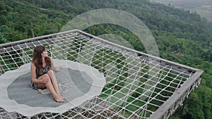 Beautiful woman sitting on a hammock net couch outdoors above green misty abyss. Korean female tourist chilling on a rug