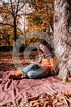 A beautiful woman sitting and leaning against a tree while reading a book in a park in a sunny autumnal day. Lifestyle autumnal
