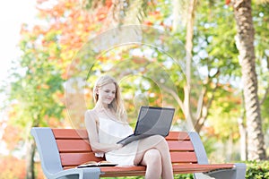 Beautiful woman sitting on a park bench using a laptop. Colorful trees in the background