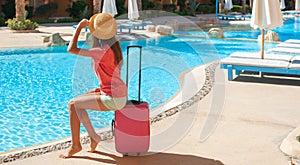 Beautiful woman sitting on pink suitcase near swimming hotel pool area. Travel, summer holidays and vacation concept