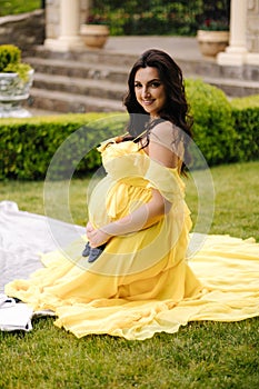 Beautiful woman is sitting on a plaid on the grass. Elegant pregnant woman waiting for baby. Future mom. Yeallow dress