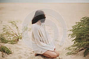 Beautiful woman sitting on sandy beach on background of green grass and looking at sea, calm tranquil moment. Stylish young female