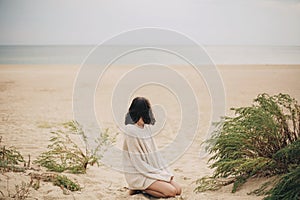 Beautiful woman sitting on sandy beach on background of green grass and looking at sea, calm tranquil moment. Stylish young female