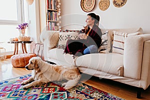 beautiful woman sitting on the sofa, enjoying a cup of tea. cute golden retriever dog besides. lifestyle indoors