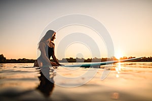 Beautiful woman sitting on surf style wakeboard on water against background of sunrise