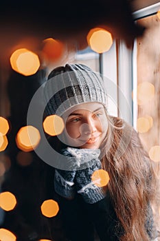 Beautiful woman sitting in a sweater home by the window. Blurred winter snow tree background. Morning, coziness, winter and people