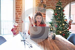 Beautiful woman sitting at the table working with laptop at home around christmas tree clapping and applauding happy and joyful,