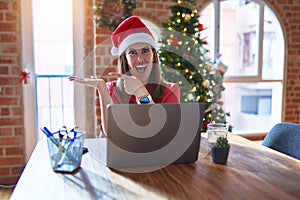 Beautiful woman sitting at the table working with laptop wearing santa claus hat at christmas amazed and smiling to the camera