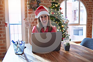 Beautiful woman sitting at the table working with laptop wearing santa claus hat at christmas with a big smile on face, pointing