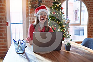 Beautiful woman sitting at the table working with laptop wearing santa claus hat at christmas happy face smiling with crossed arms