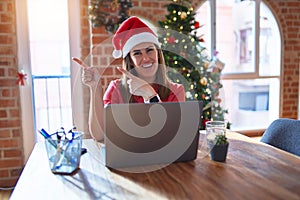 Beautiful woman sitting at the table working with laptop wearing santa claus hat at christmas smiling and looking at the camera