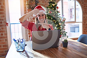 Beautiful woman sitting at the table working with laptop wearing santa claus hat at christmas smiling making frame with hands and