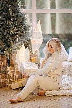 Beautiful woman sitting on windowsill by Christmas tree. Blond hair woman in white costume have rest at home. Christmas