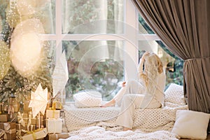 Beautiful woman sitting on windowsill by Christmas tree. Blond hair woman in white costume have rest at home. Christmas