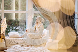 Beautiful woman sitting on windowsill by Christmas tree. Blond hair woman in white costume have rest at home. Christmas