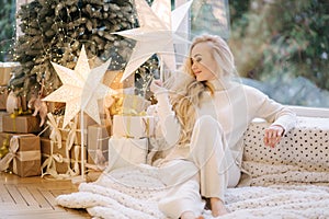 Beautiful woman sitting on windowsill by Christmas tree. Blond hair woman in white costume have rest at home. Christmas