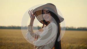 Beautiful woman smiles standing alone in straw hat standing alone at sunset near agronomic rural field with crops