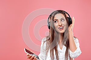 Beautiful woman smiling and enjoying favorite music, via wireless headphones over pink background.