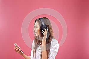 Beautiful woman  smiling looking aside with smartphone in hands enjoying favorite music, via wireless headphones over pink backgro