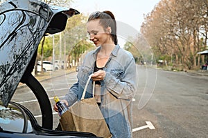 Beautiful woman in sportswear standing near the car, putting bottle of water into handbag while going to gym for