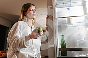 Beautiful woman standing on kitchen at night and looking in open fridge holding green apple in hand