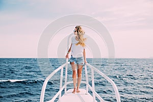 Beautiful woman standing on the nose of the yacht at a sunny summer day, breeze developing hair, beautiful sea on background