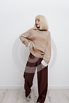 a beautiful woman stands half sideways in full height on a light background in a sweater and shirt, putting her hand on