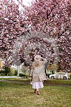 Beautiful woman stands with her back near the sakura trees. Woman in hat, dress and stylish coat. Pink flowers blooming in