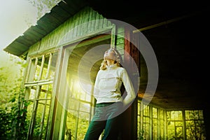 A beautiful woman stands near the broken windows of an old wooden house on a sunny warm summer day. Youth vibe