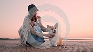 Beautiful woman stroking corgi dog while sitting on sea beach on evening.