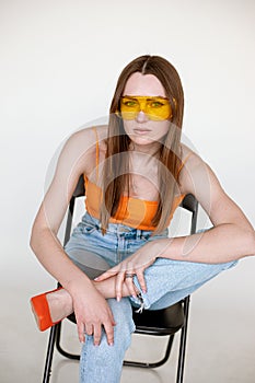 Beautiful woman in stylish sunglasses sitting on black chair and wearing seasonal blue denim and bright orange top