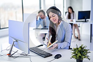 Beautiful woman in suit and headset opening book to check notes while talking to a costumer at call center.