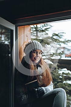 Beautiful woman in a sweater  sitting home by the window. Blurred winter snow tree background. Morning, coziness, winter and peopl