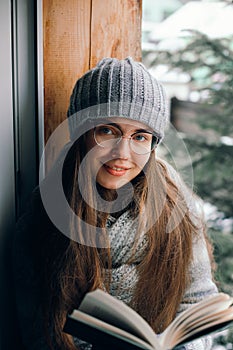 Beautiful woman in a sweater  sitting home by the window. Blurred winter snow tree background. Morning, coziness, winter and peopl