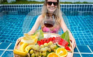 Beautiful woman in the swimming pool with glass of wine and floating tray of fruits