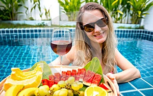 Beautiful woman in the swimming pool with glass of wine and floating tray of fruits